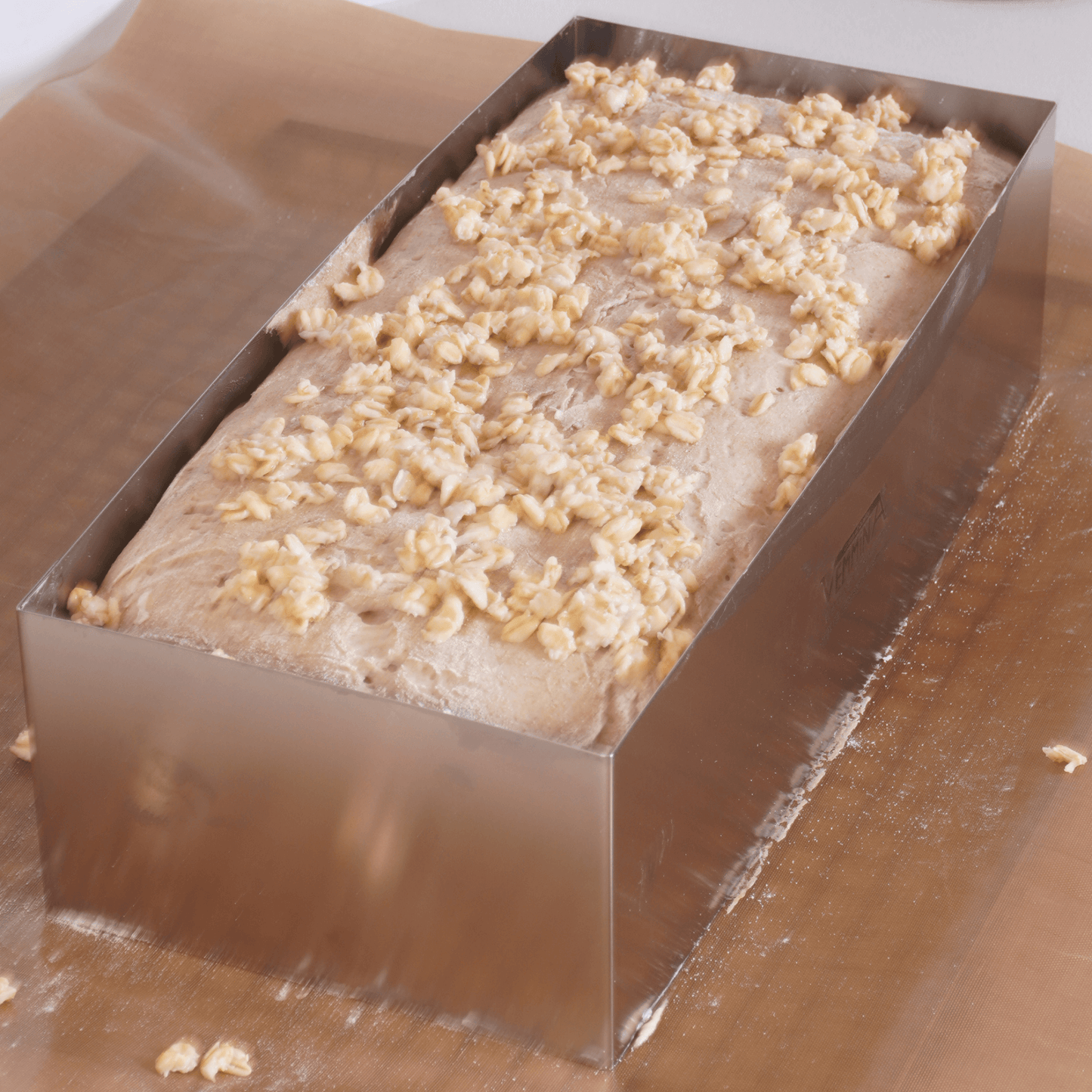 Freshly prepared dough topped with oats in a metal bread baking mold on parchment paper.